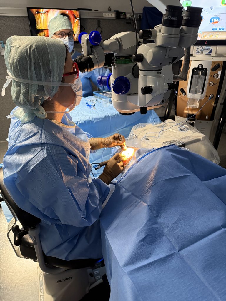 Le Dr Alina Stalker au microscope opératoire Carl Zeiss pendant une opération de la cataracte, instrument chirurgical en main, champ stérile en place.